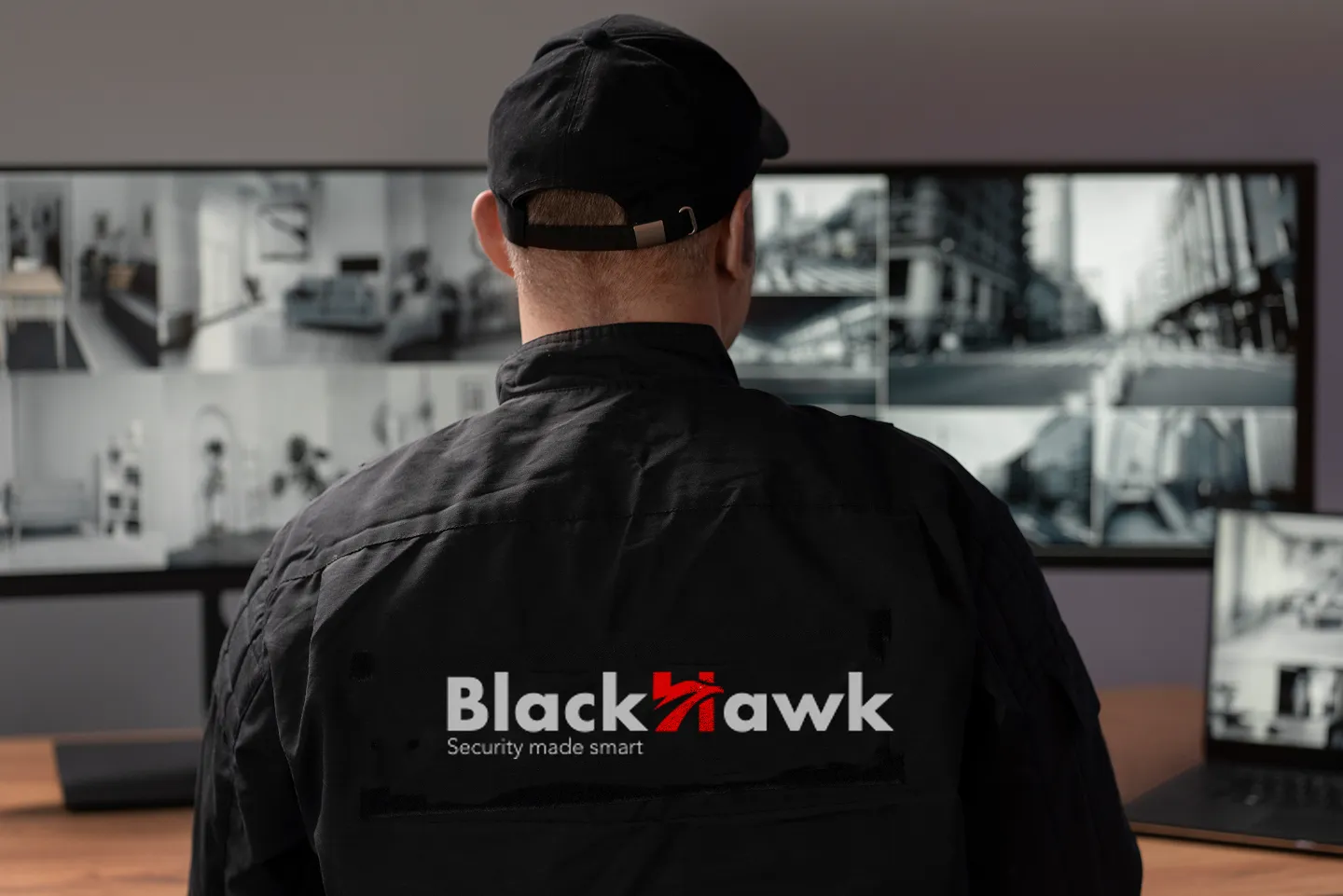 Security guard wearing a Blackhawk jacket monitoring multiple surveillance screens.