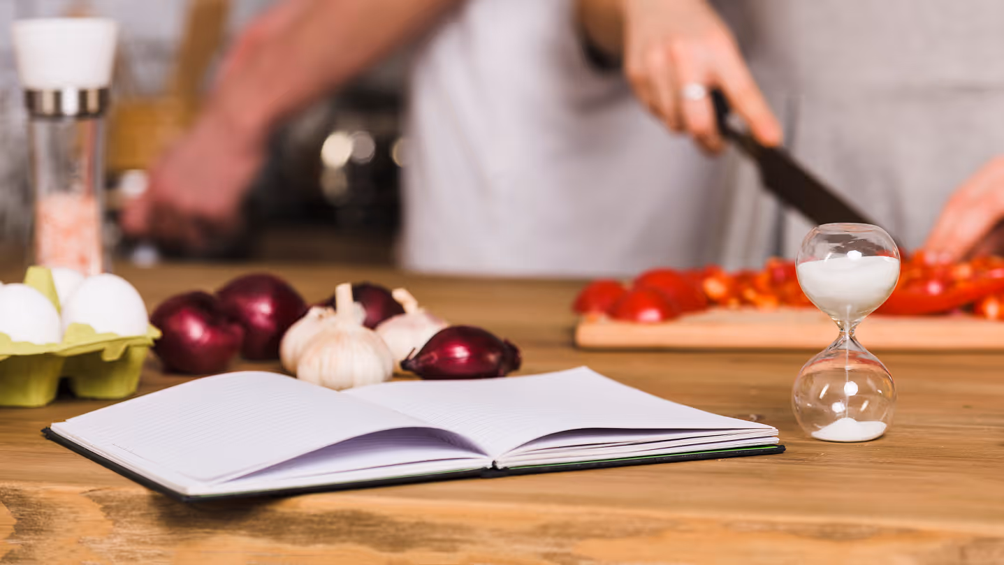 Open blank notebook with eggs, onions, garlic, and an hourglass on a wooden table while someone chops tomatoes.