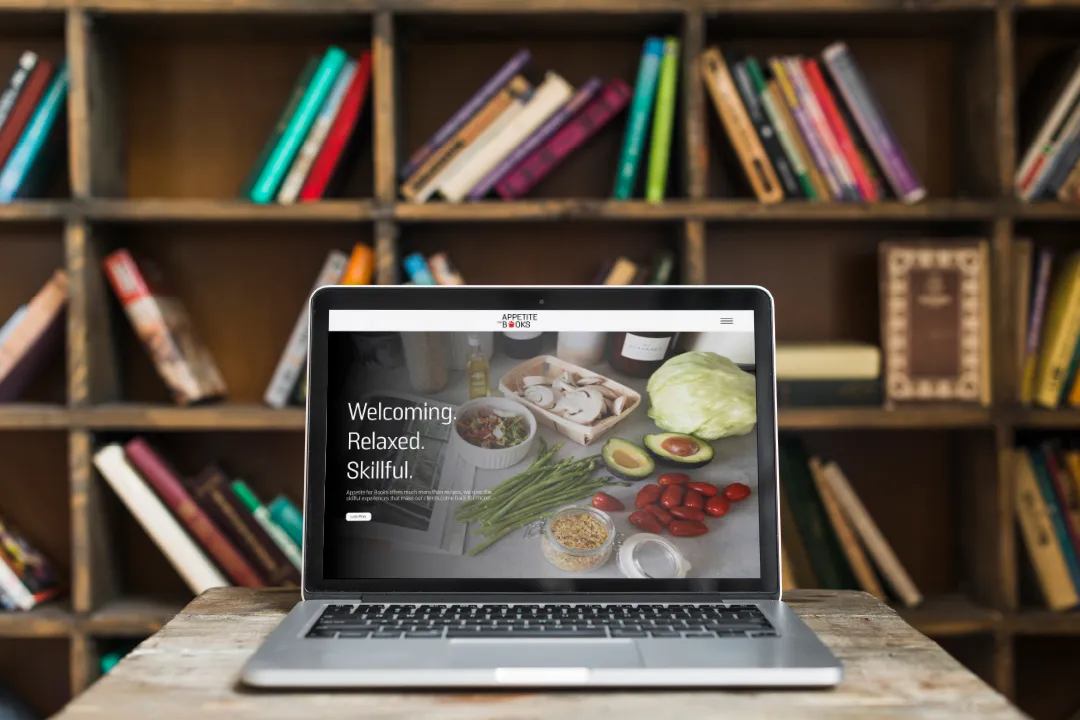 Laptop on wooden table displaying fresh vegetables and words 'Welcoming. Relaxed. Skillful.' with bookshelf background.