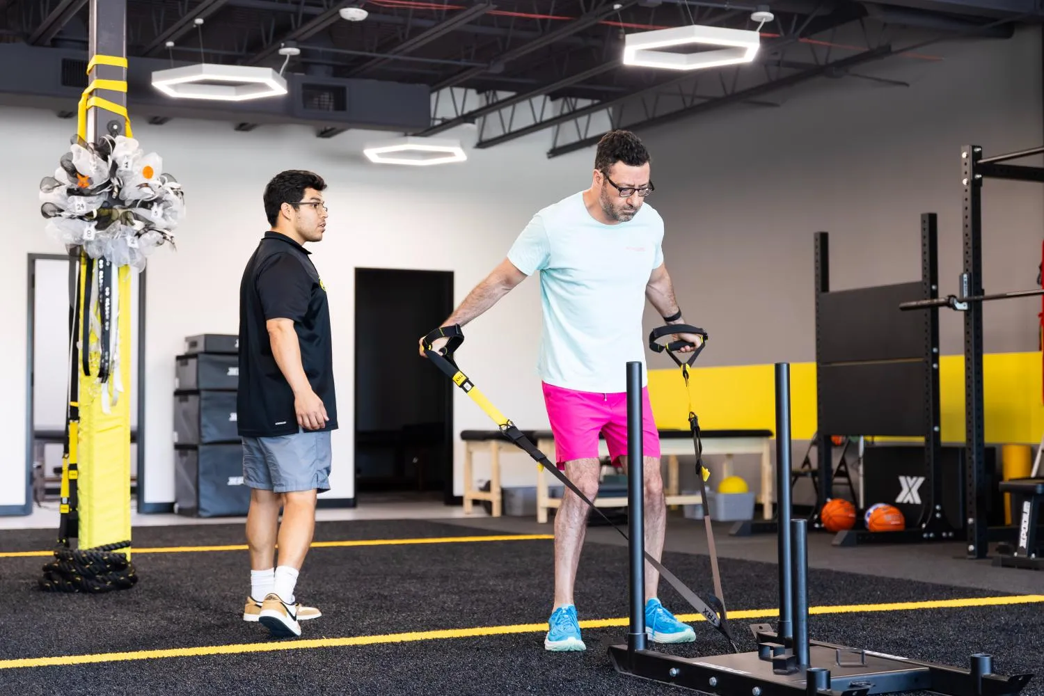 Man in pink shorts exercising with resistance straps on sled while trainer watches in gym.