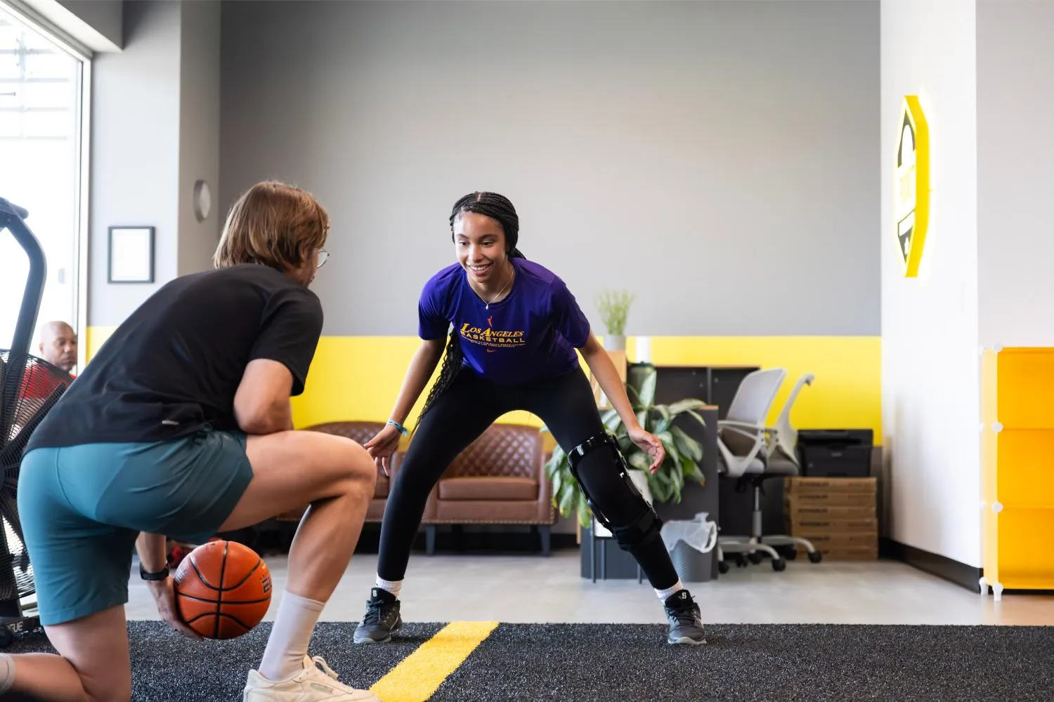Young woman in purple shirt with leg brace practicing basketball defense against coach indoors.