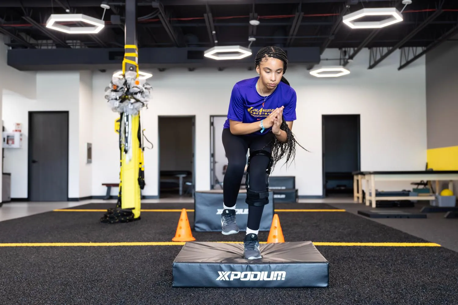 Athlete with knee brace doing a step-up exercise on a padded platform in a gym.