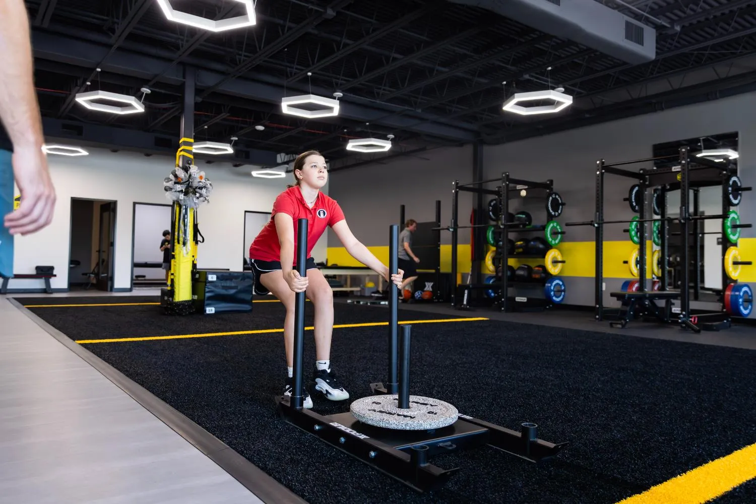 Young woman in a red shirt pushing weighted sled in a modern gym with black and yellow decor.
