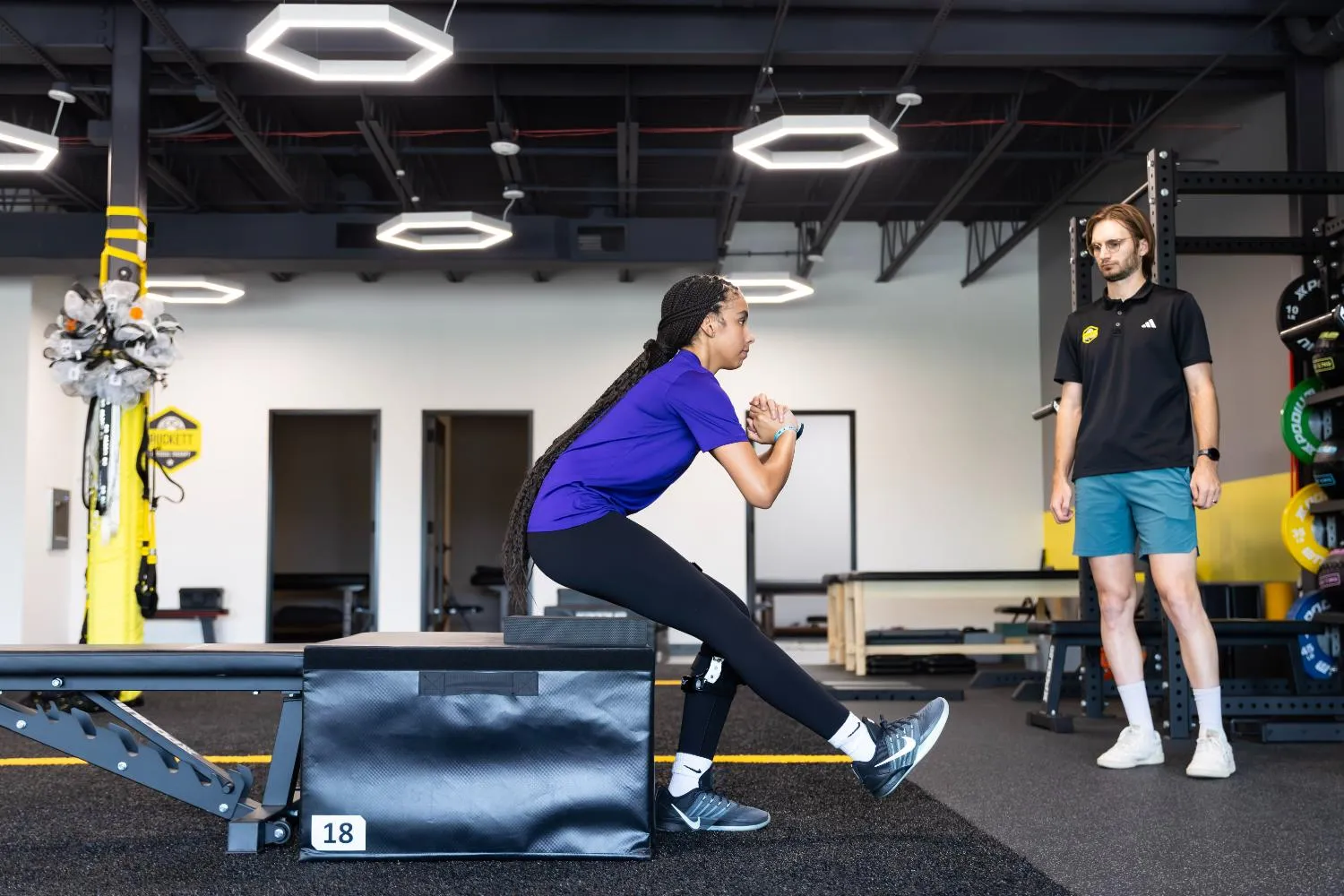 Person with prosthetic leg doing seated leg stretch while trainer watches in gym.