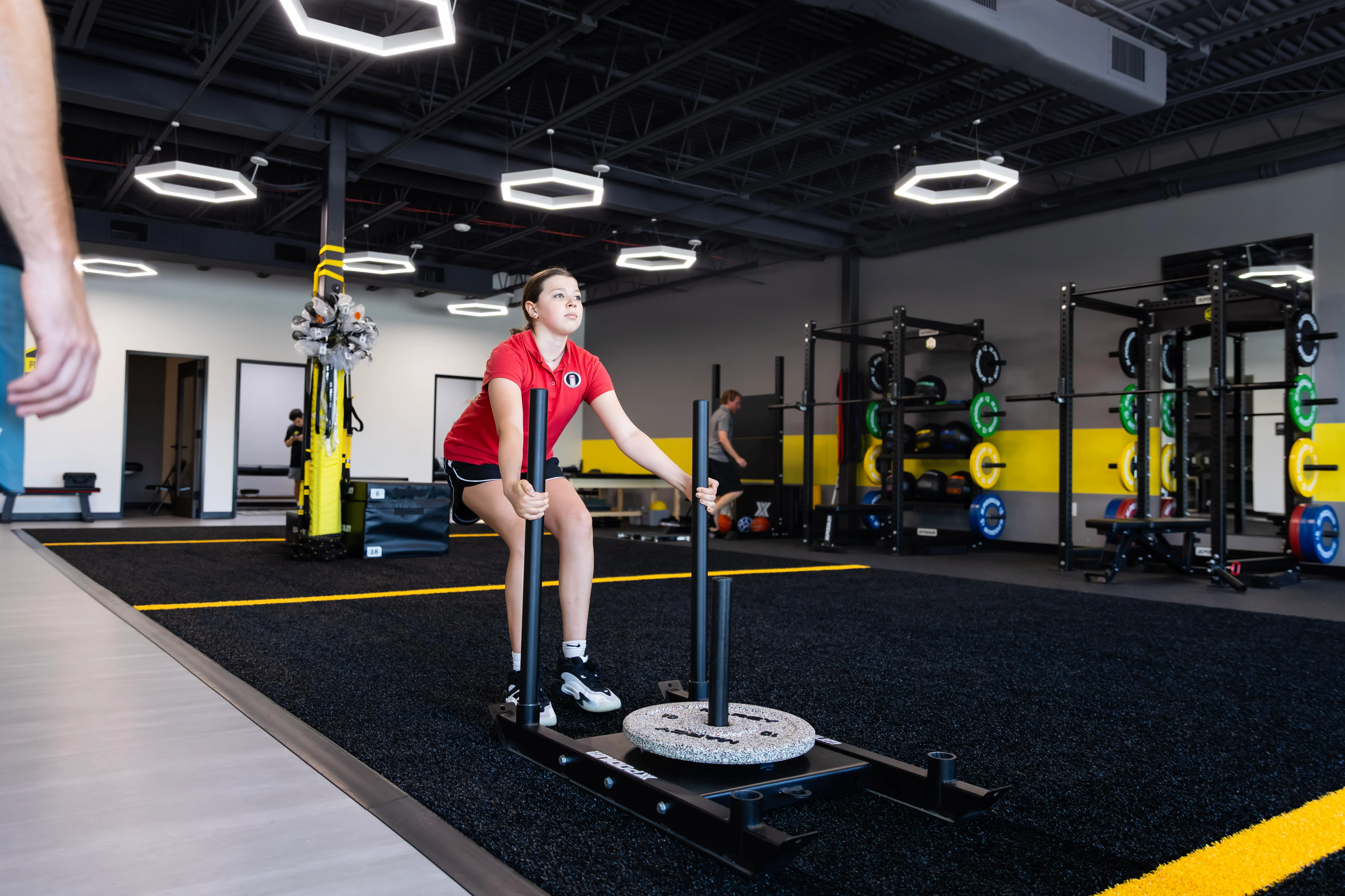 Young girl in red shirt pushing weighted sled during workout in modern gym.
