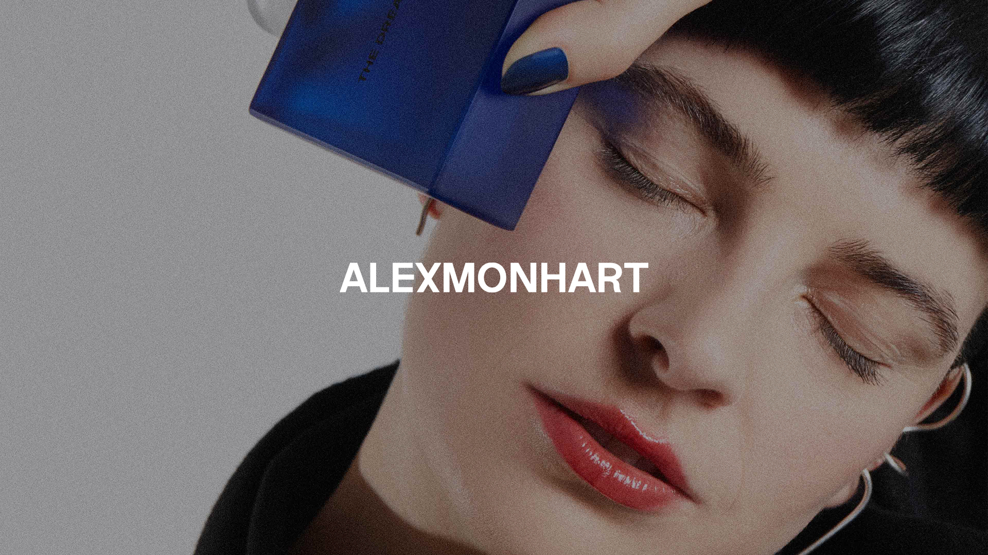 Close-up of a woman with red lips and blue nail polish holding a blue bottle near her closed eye.