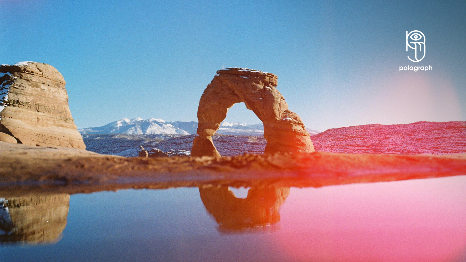 Natural sandstone arch with snowy mountains reflected in calm water under clear blue sky.