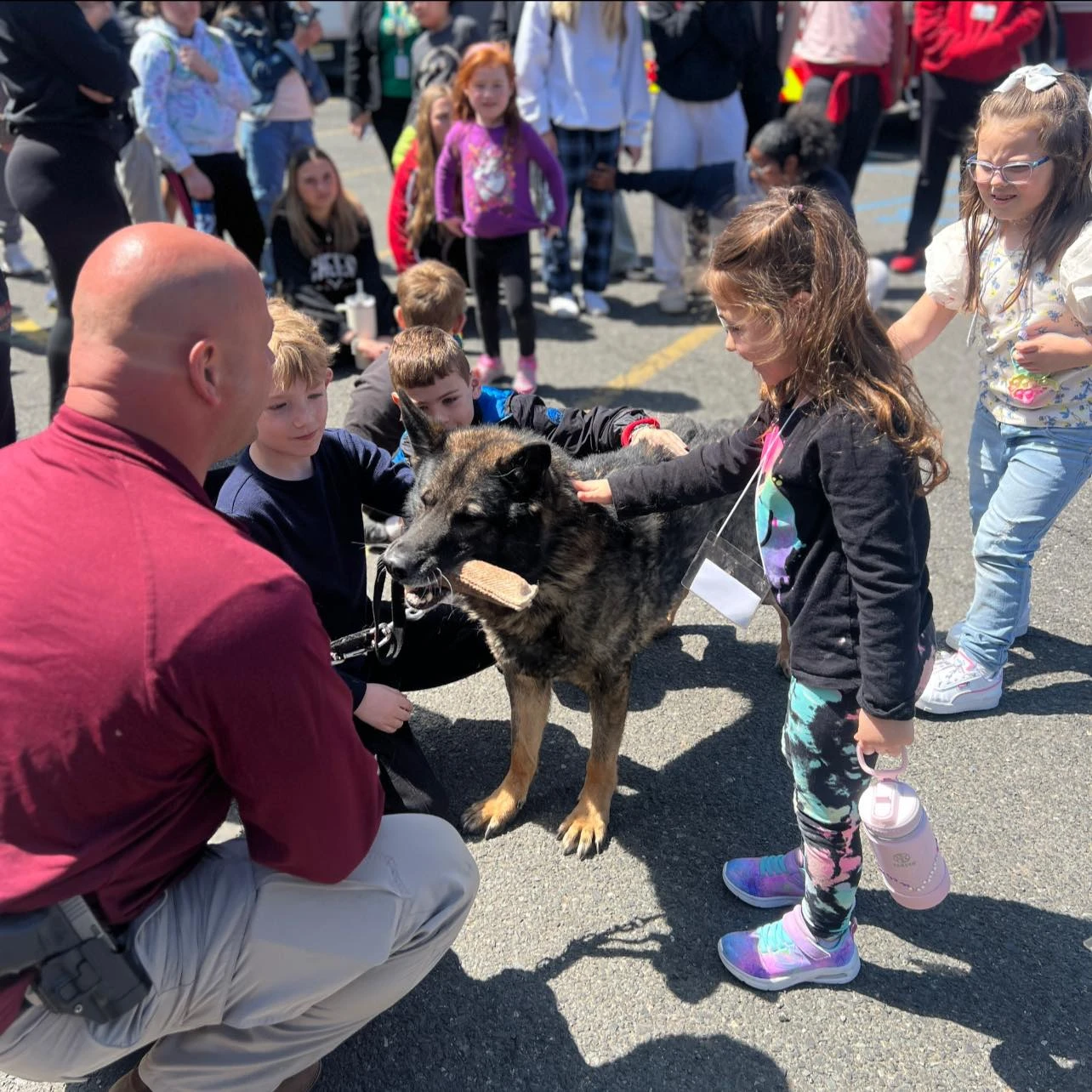 Children petting a German Shepherd holding a training bite sleeve, with a man kneeling nearby outdoors.