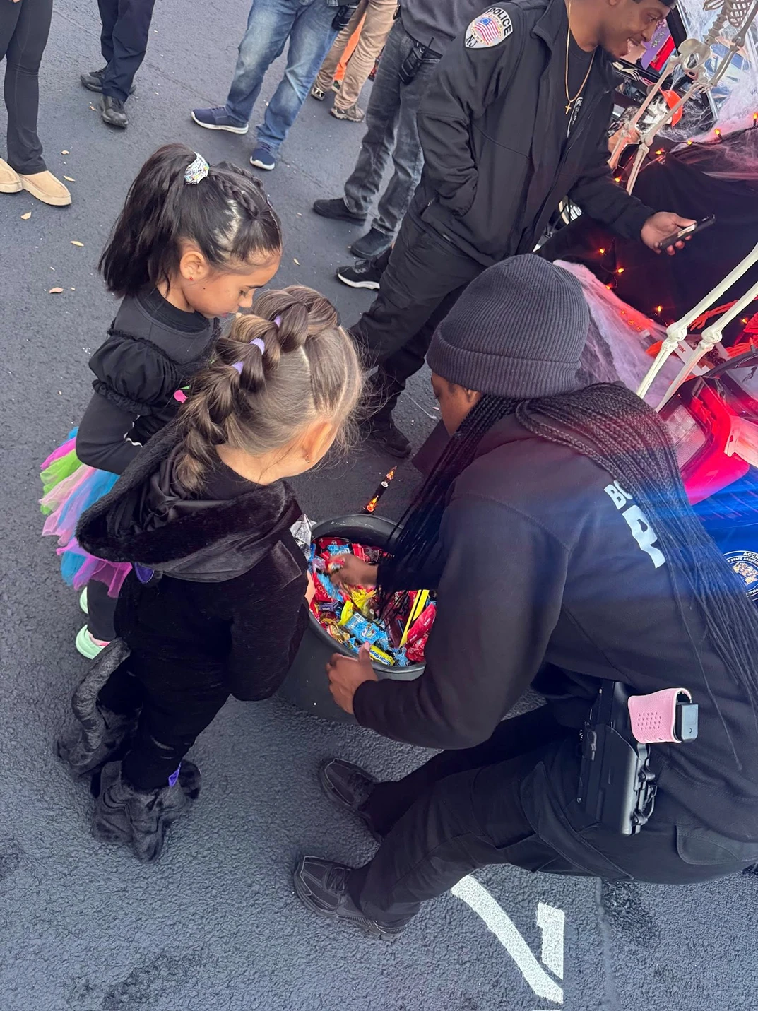 Two girls in Halloween costumes receiving candy from a person in black near a police officer.