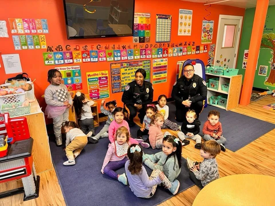 Two police officers with a group of young children sitting and standing in a colorful classroom.