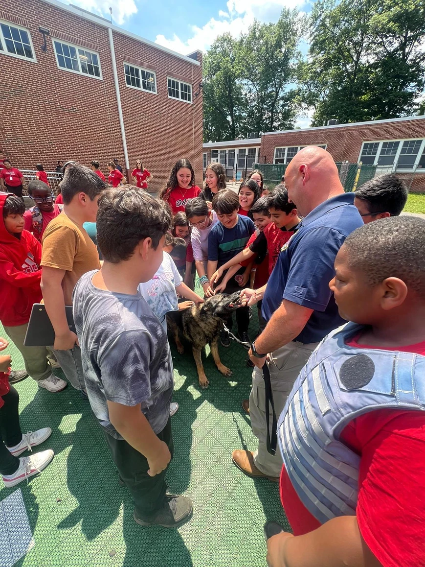 Group of children gathered around a man with a police dog outdoors on a sunny day.