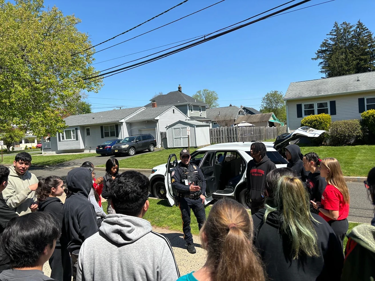 Police officer talks to a group of diverse teenagers outside on a sunny neighborhood street.