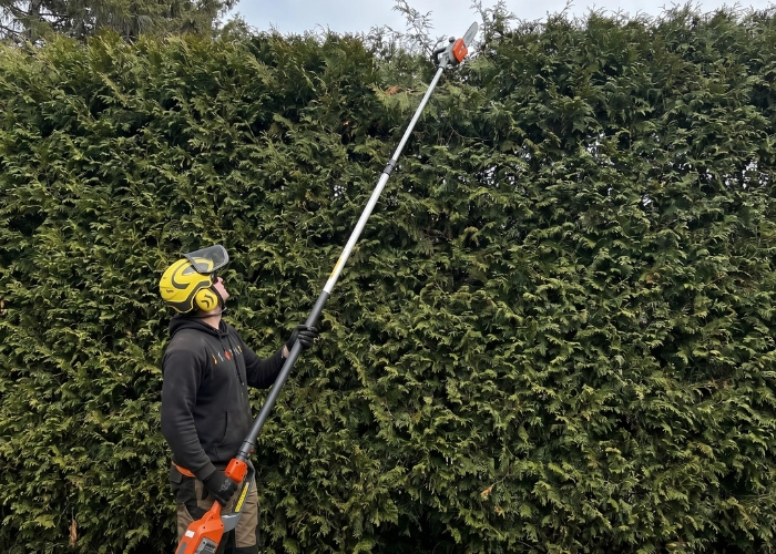 Worker using a pole hedge trimmer to shape the top of a tall cedar hedge during hedge trimming in Montreal