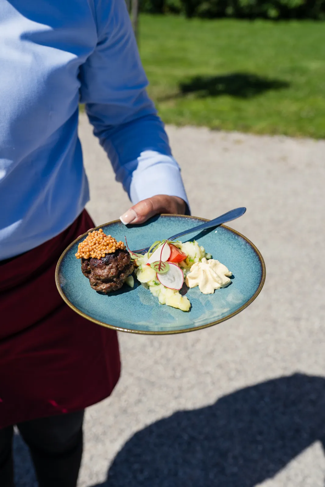 Person hält blauen Teller mit gebratenem Hackfleisch, Kartoffelsalat, Senf und Gabel im Freien.