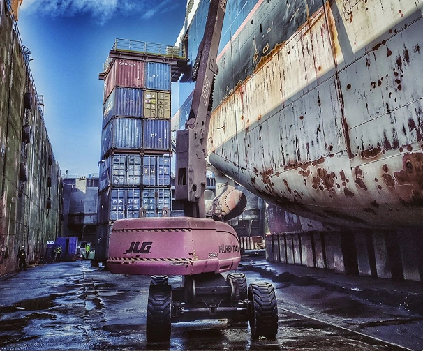 Grue élévatrice rouge entre conteneurs empilés et coque de navire rouillée dans un chantier naval.