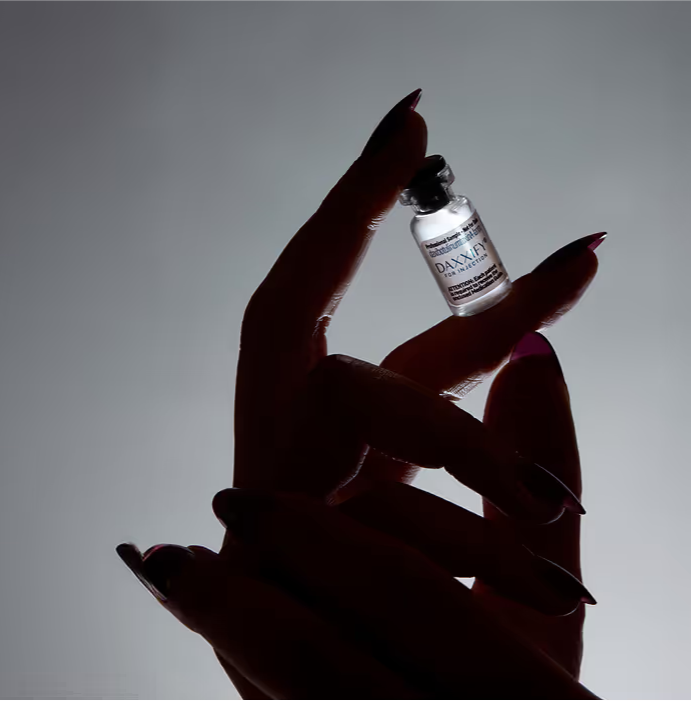 Hand with long nails holding a small vial labeled DAXIVI, backlit on gray background.