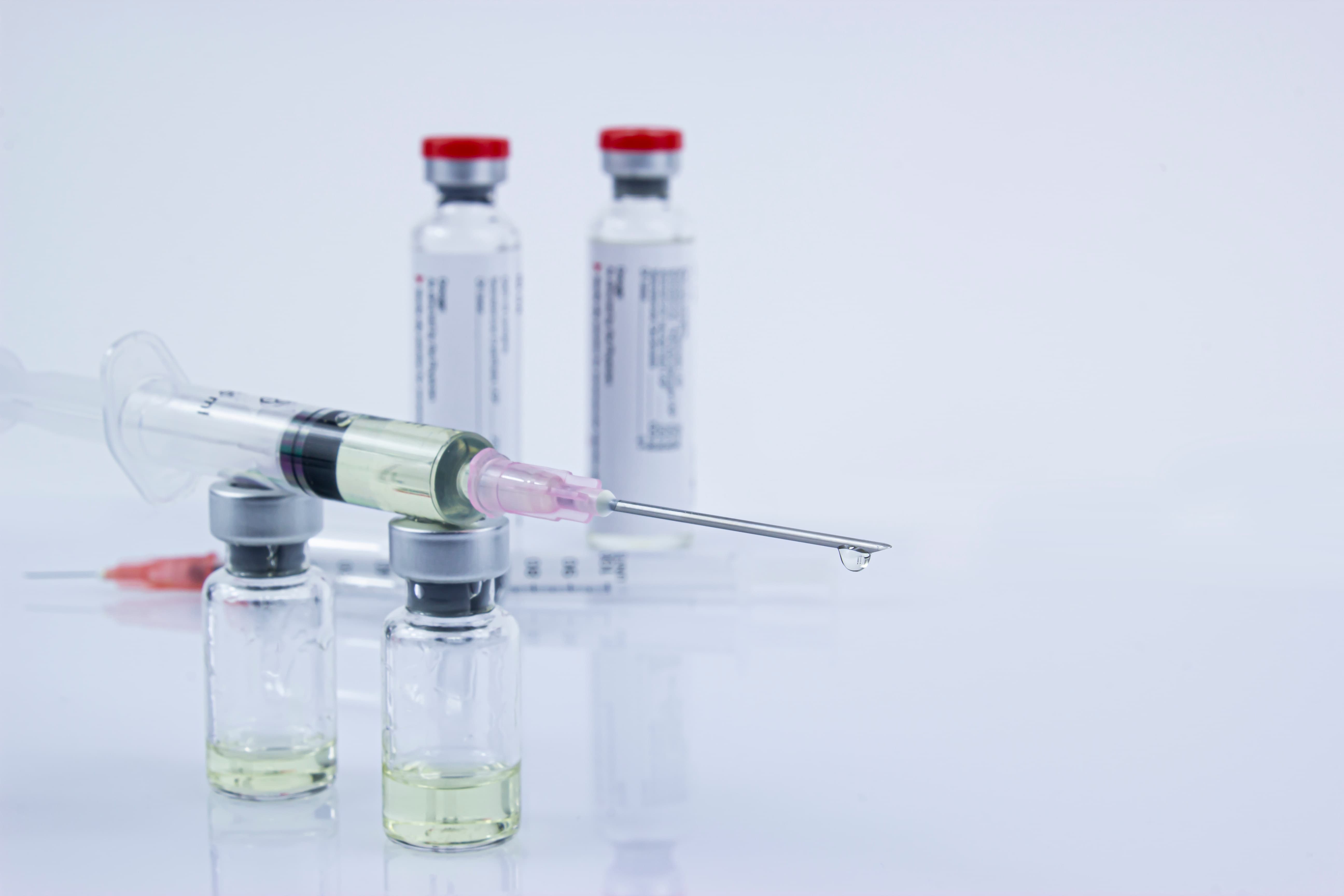 Syringe with liquid and a droplet at the needle tip resting on two vials with liquid inside.