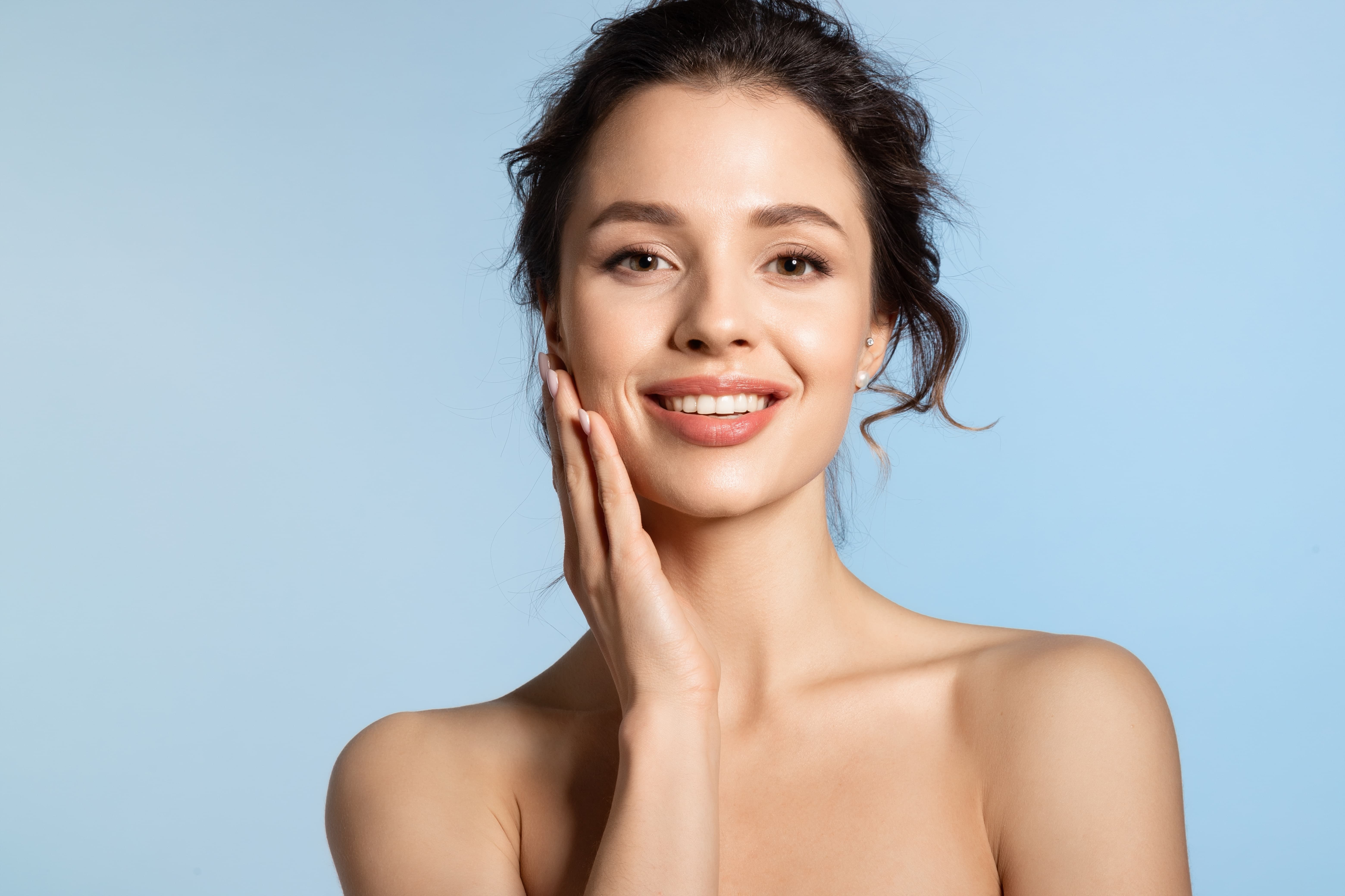 Smiling woman with clear skin touching her face against a light blue background.