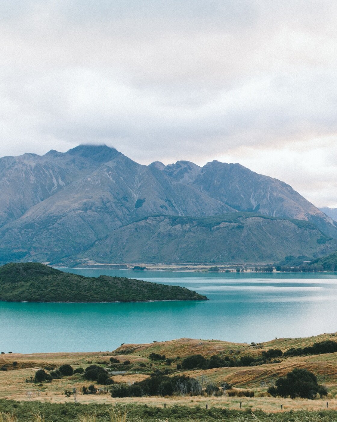 Calm turquoise lake with green islands and a range of tall, rocky mountains under a cloudy sky.