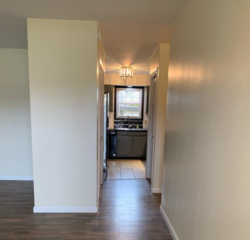 Narrow hallway with light beige walls and wood flooring leading to a kitchen with a window and overhead light.