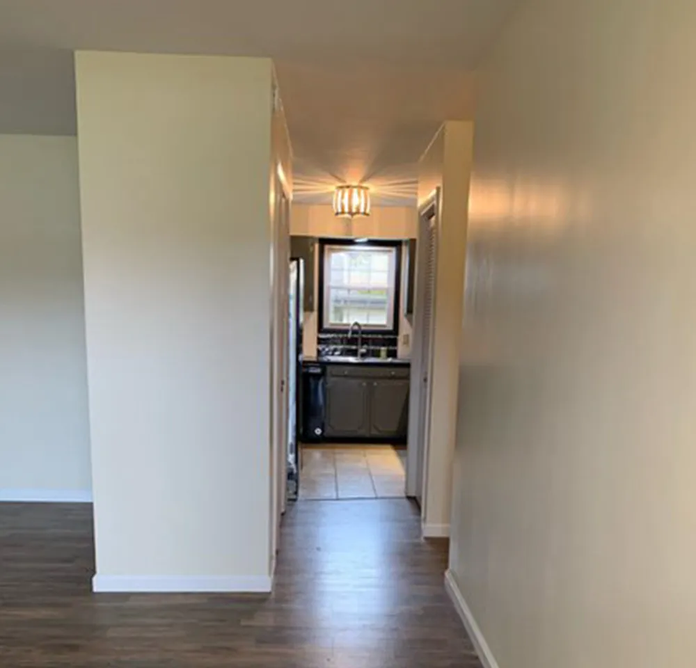 Narrow hallway with light beige walls and wood flooring leading to a kitchen with a window and overhead light.