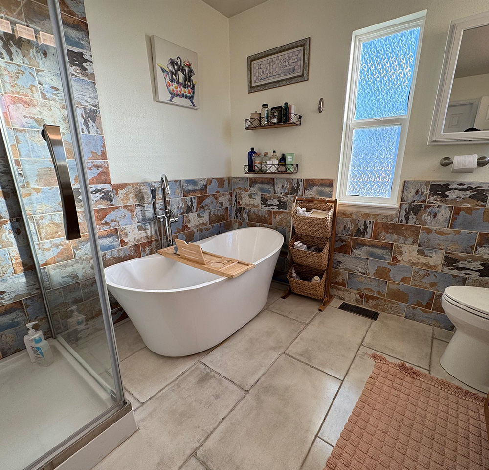 Bathroom with white freestanding tub, stone tiles on walls, glass shower door, and woven storage baskets.
