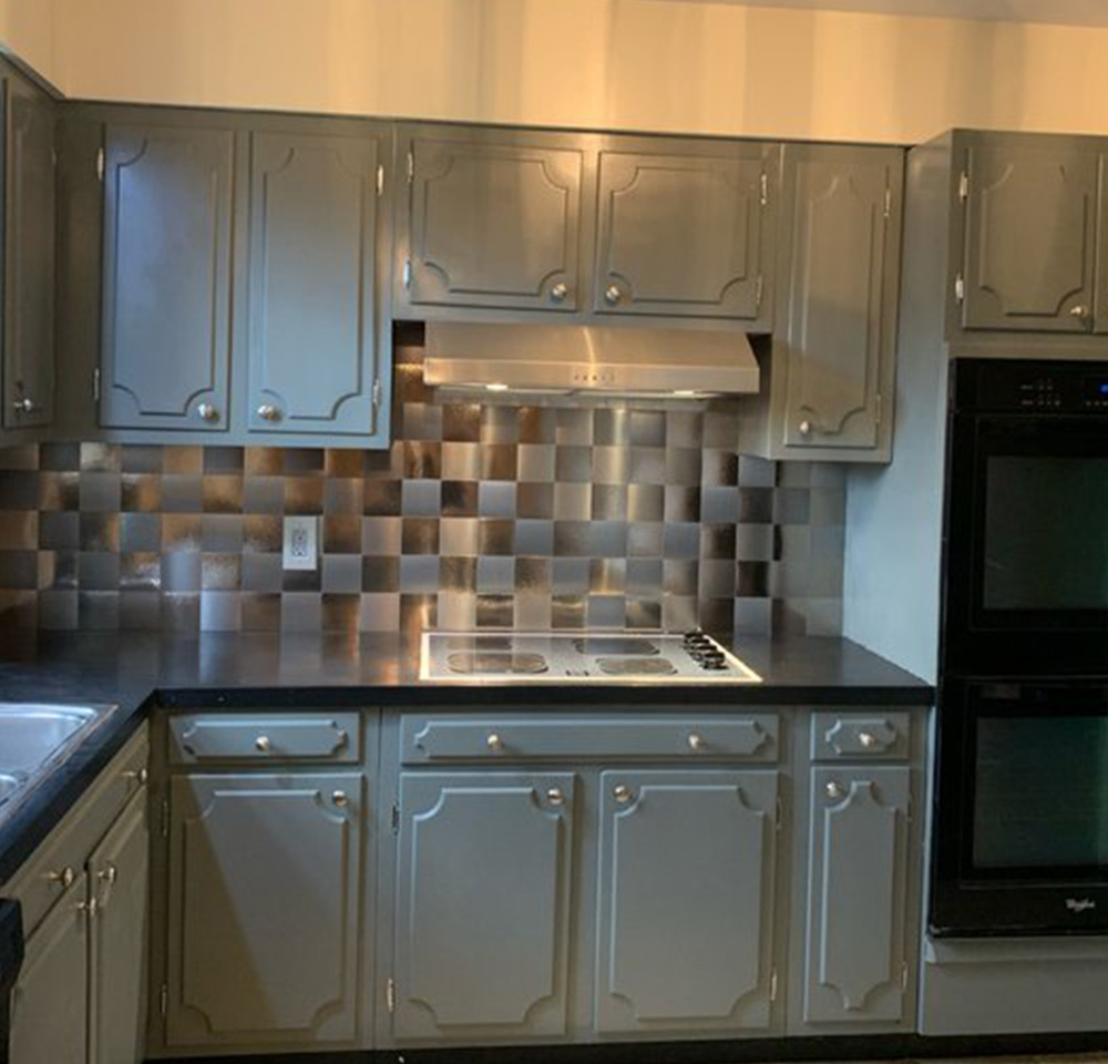 Kitchen with gray cabinets, black countertop, stainless steel stove and hood, and metallic tile backsplash.
