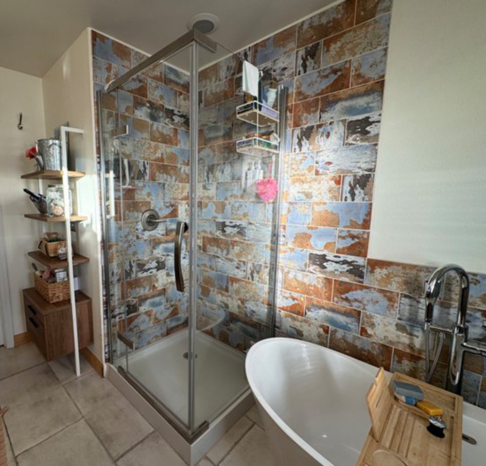 Bathroom with rustic multicolored tile shower, glass door, white bathtub, and wooden storage shelves.