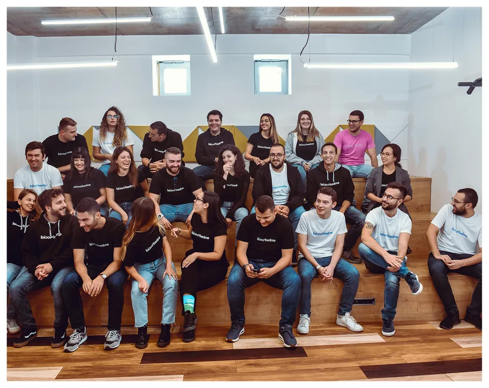 Group of twenty diverse young adults sitting on wooden tiered seating, wearing black and white company logo shirts, smiling and interacting in a modern indoor space.