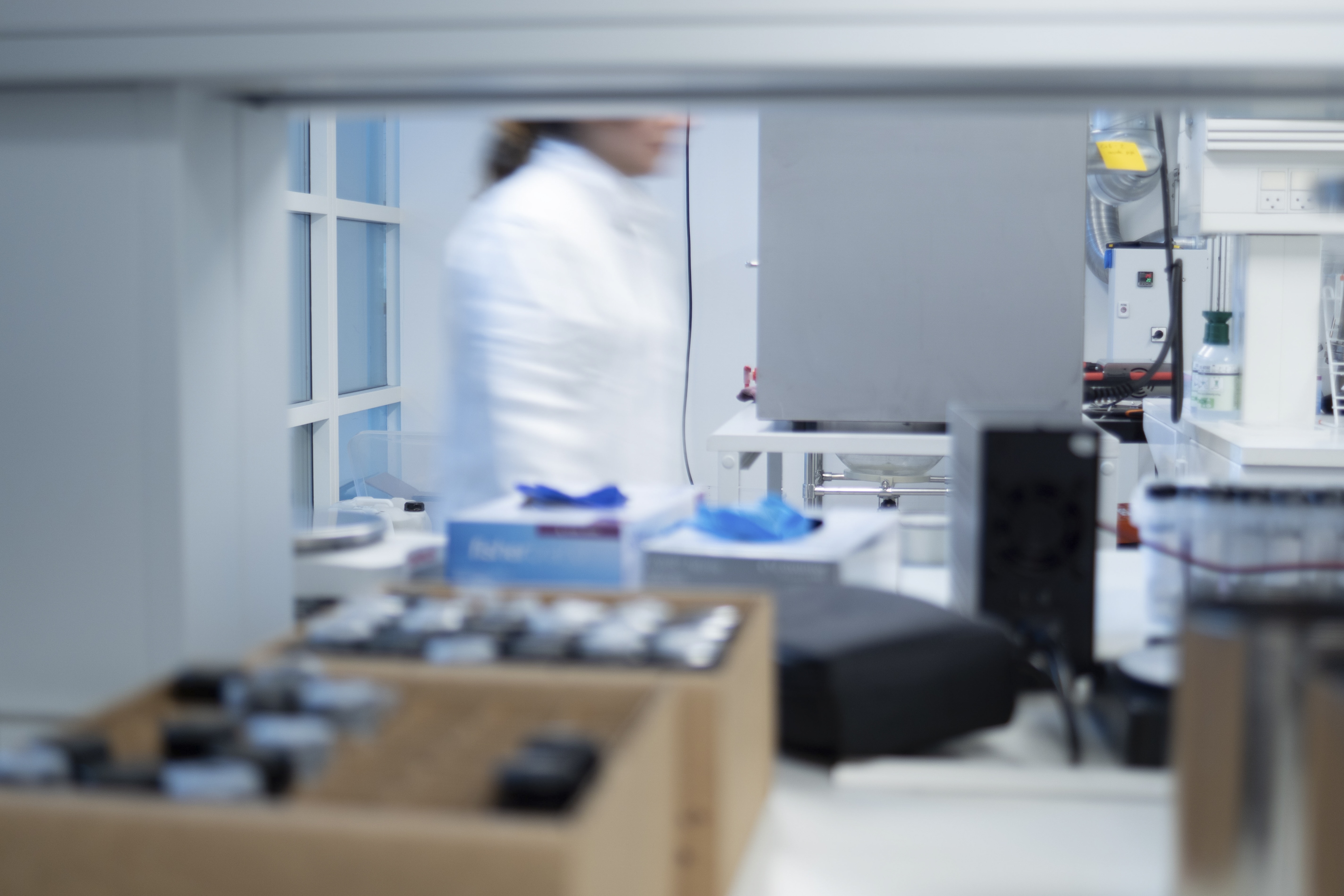Scientist blurred in background of a lab with equipment and samples on a white bench.