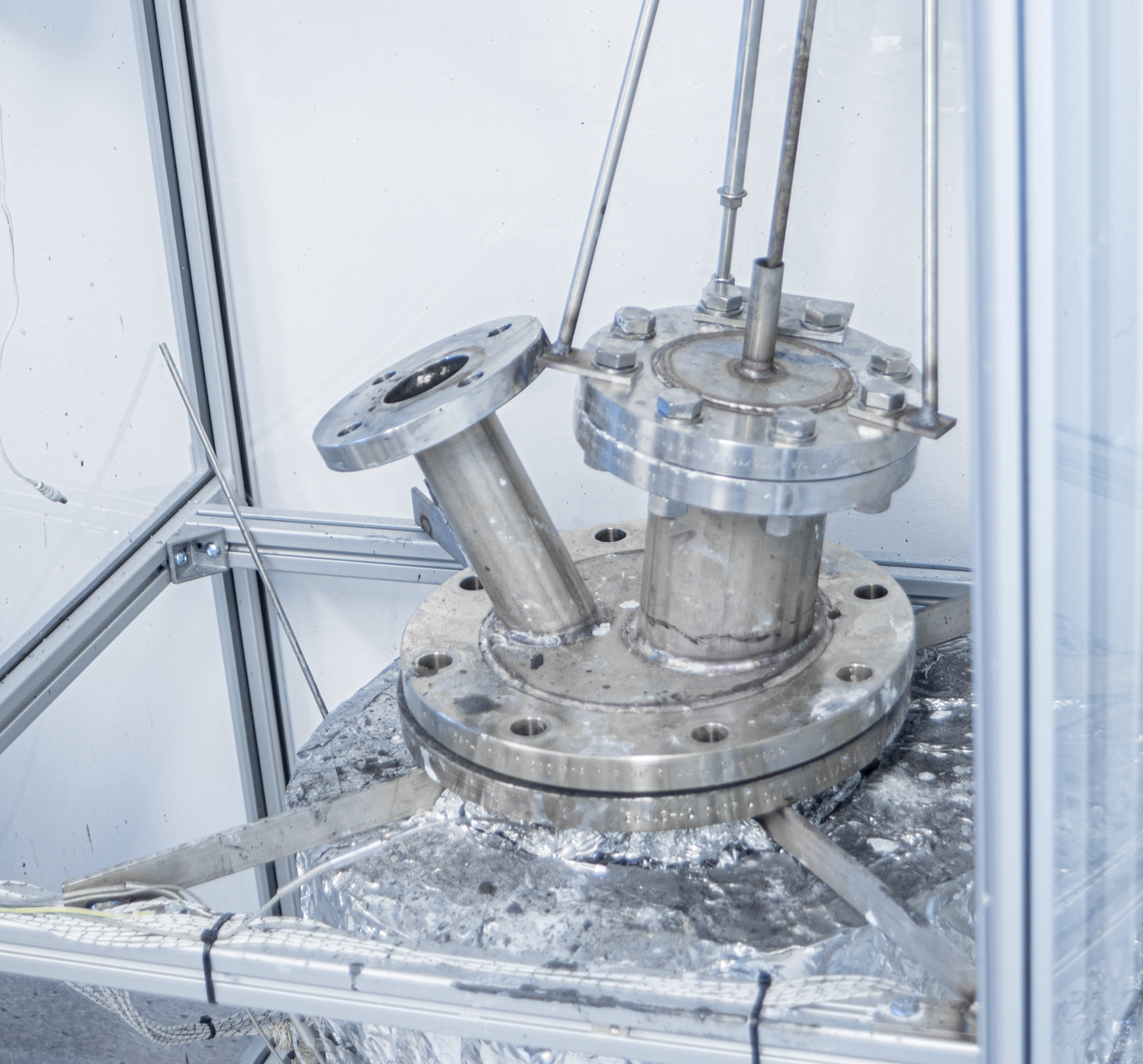 Metal industrial reactor vessel with flanges and pipes inside a protective enclosure.