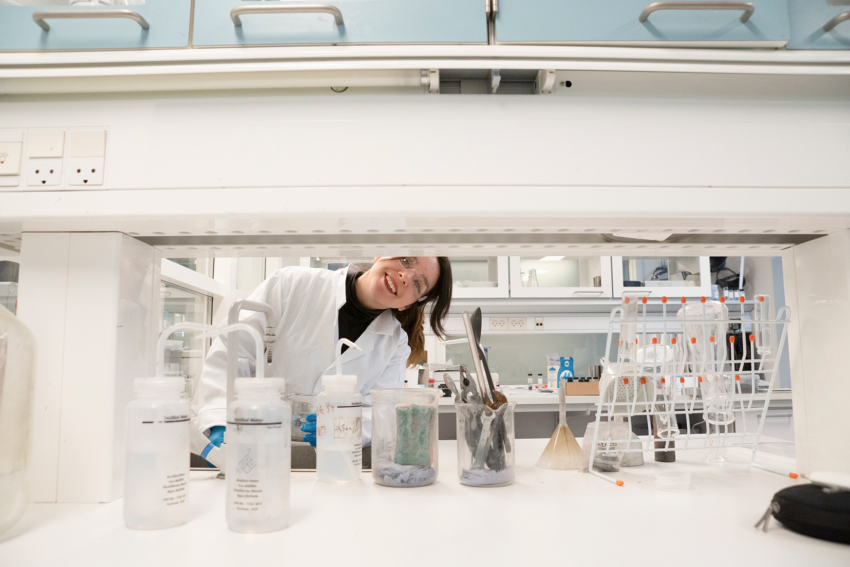Smiling scientist in a white lab coat working in a laboratory with glassware and tools on the counter.