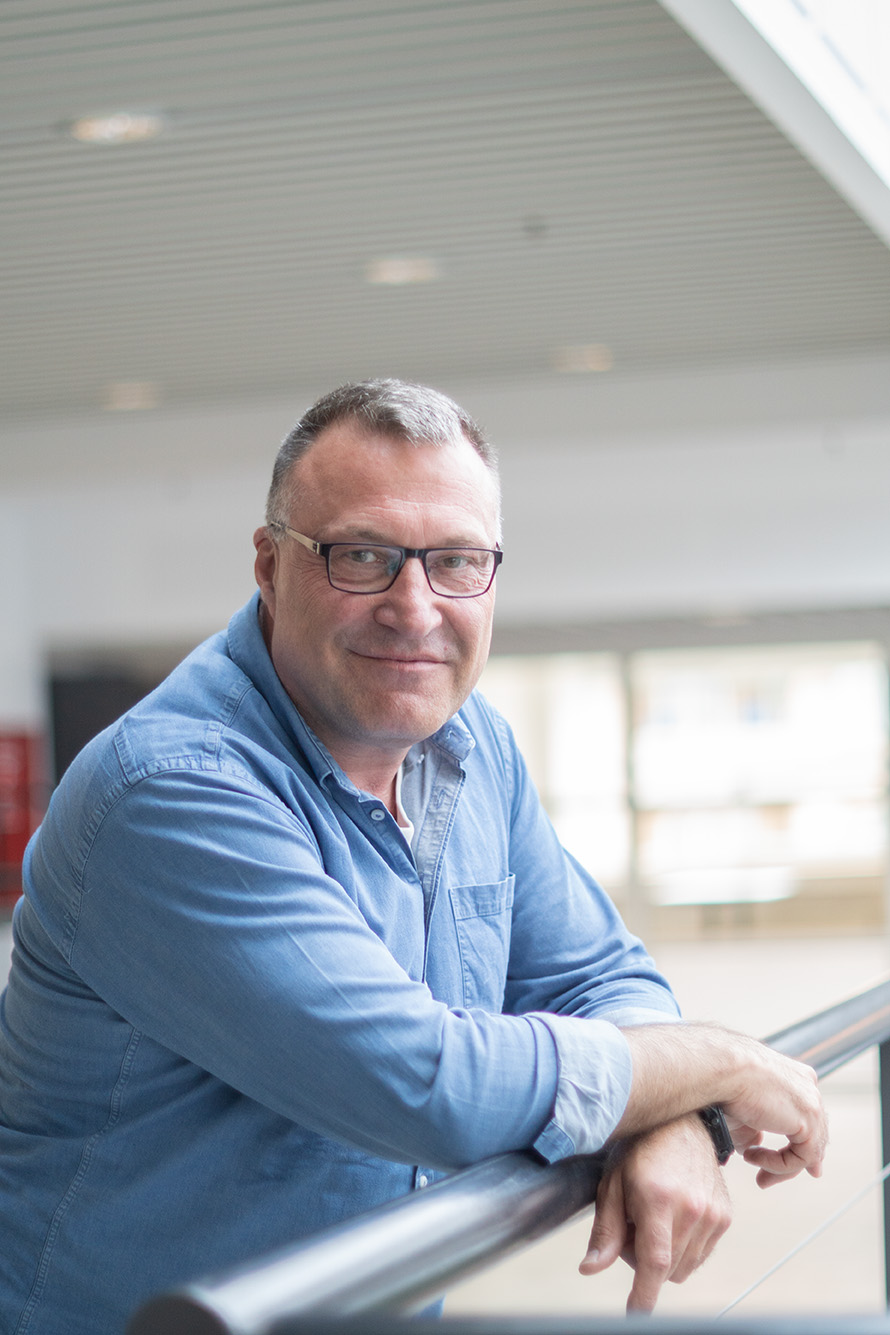 Middle-aged man in glasses and blue shirt leaning on railing indoors, smiling at the camera.