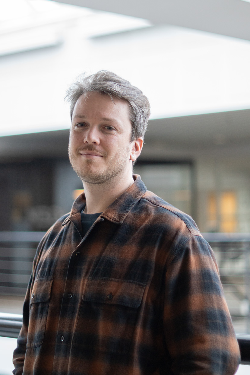 Man with light beard wearing a brown and black plaid shirt, standing indoors near a railing.