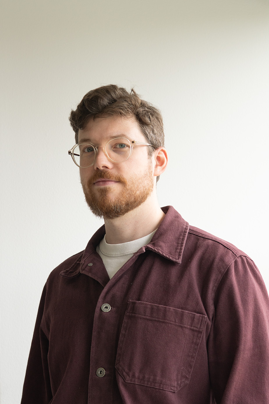 Man with red beard and glasses wearing a maroon jacket and white shirt against a plain background.