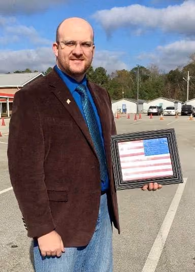 Man in glasses and brown jacket holding framed American flag with message in a parking lot.