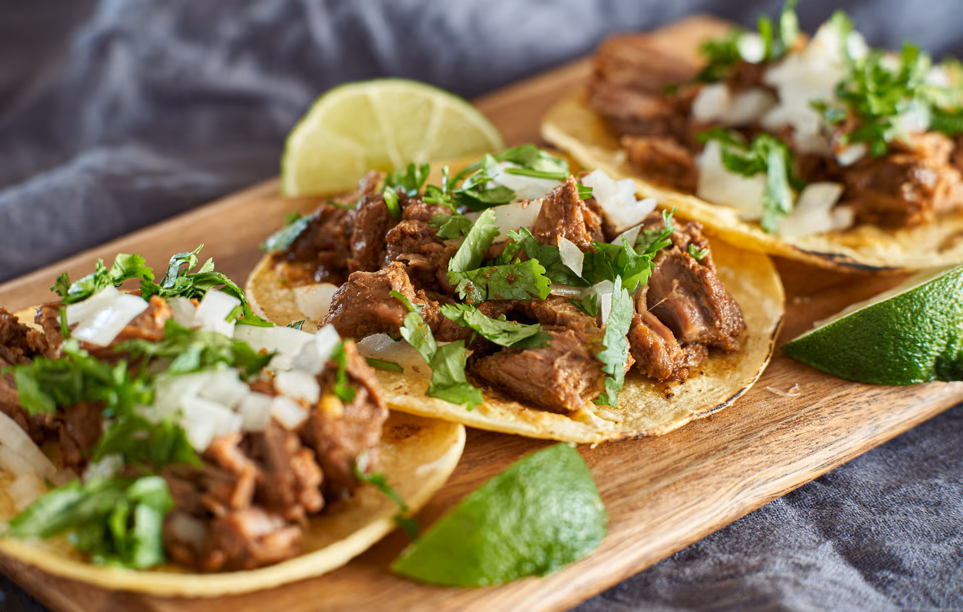 Three tacos with seasoned meat, chopped onions, cilantro, and lime wedges on a wooden board.