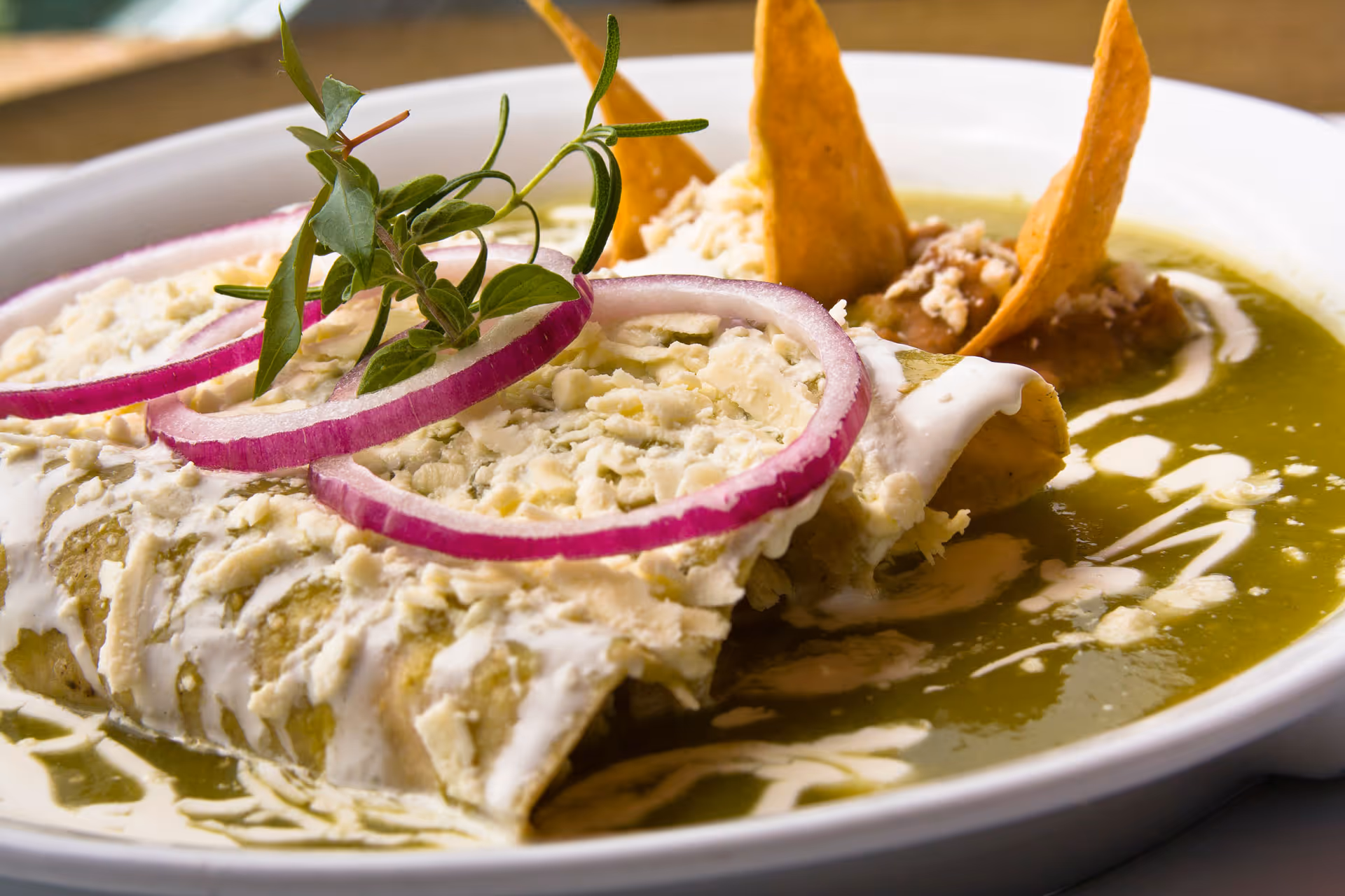 Enchiladas topped with cream, cheese, onion rings, and herbs served with chips and beans in green sauce.
