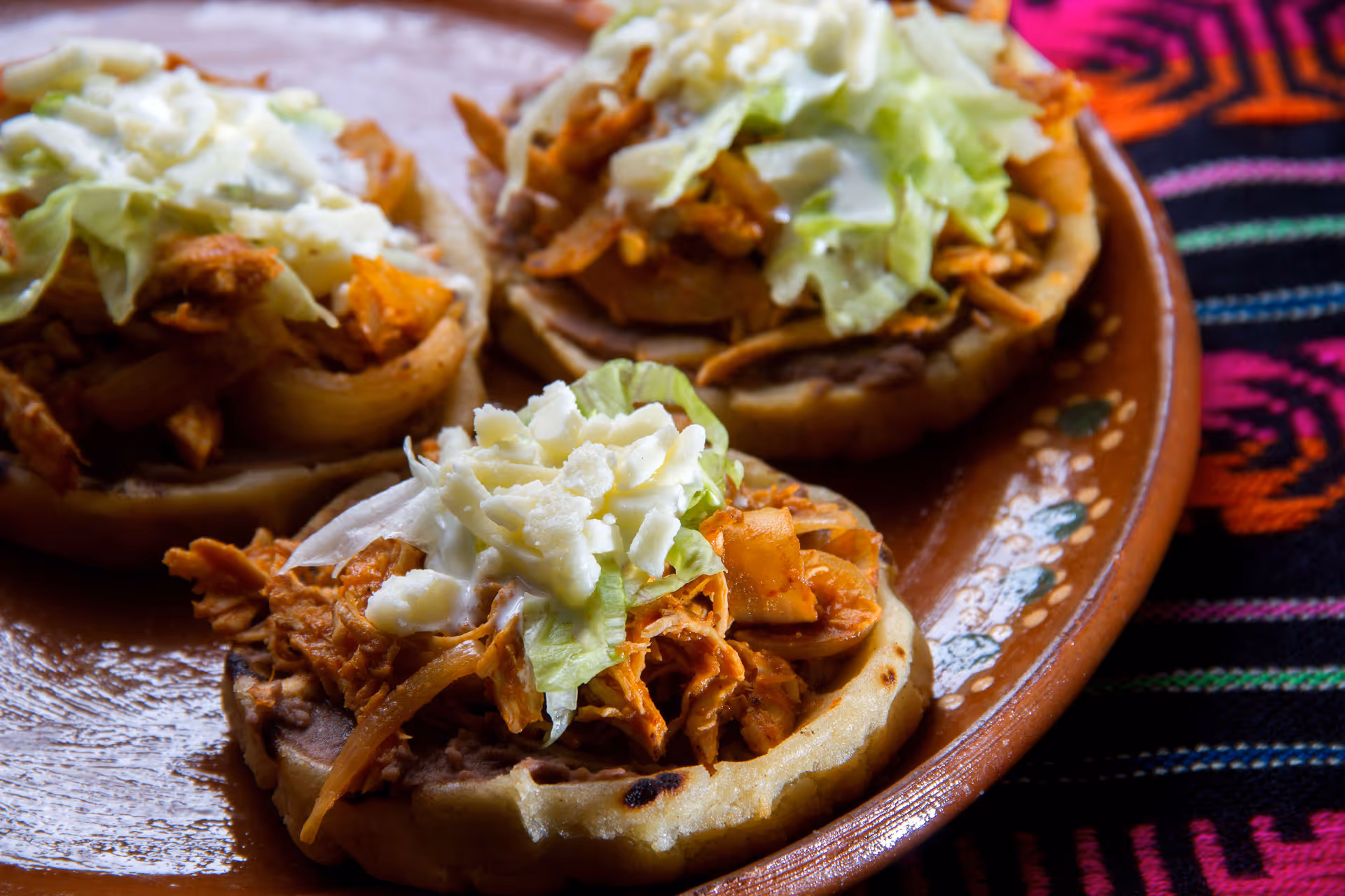 Three sope Mexican dishes with shredded chicken, lettuce, cheese, and refried beans on a brown plate.