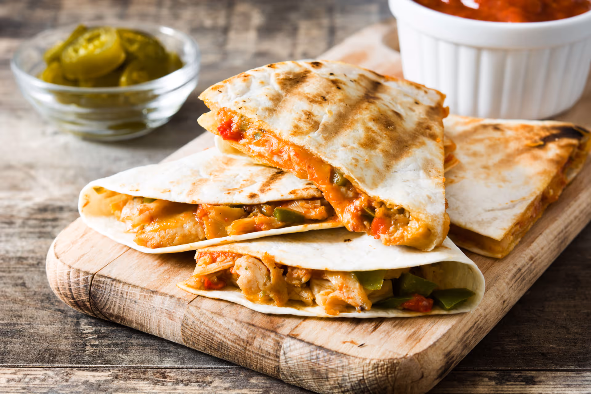 Grilled chicken quesadilla with peppers on wooden board with jalapeños and salsa in background.