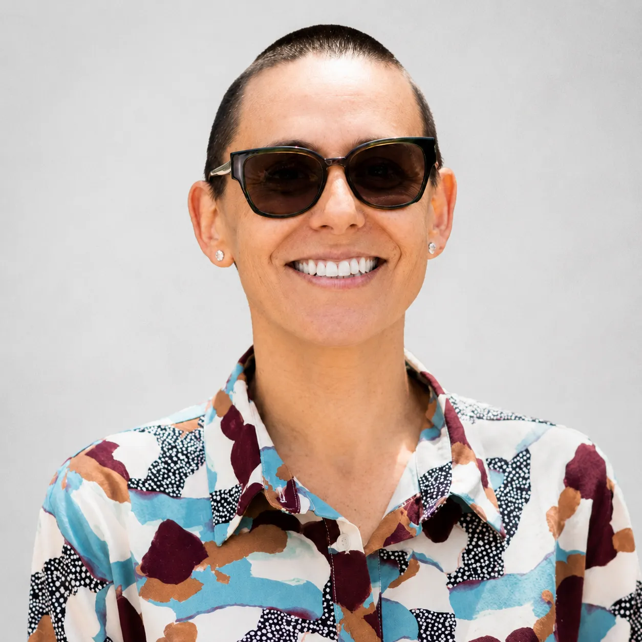 Person with short hair and sunglasses smiling, wearing a patterned shirt with maroon, blue, white, and tan colors.