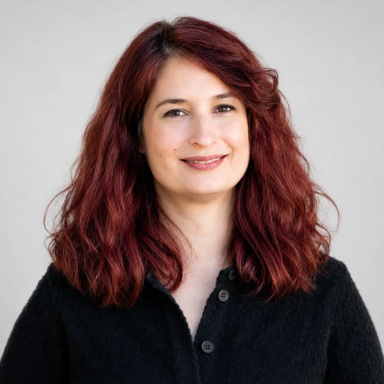 Smiling woman with shoulder-length wavy red hair wearing a black button-up sweater against a gray background.