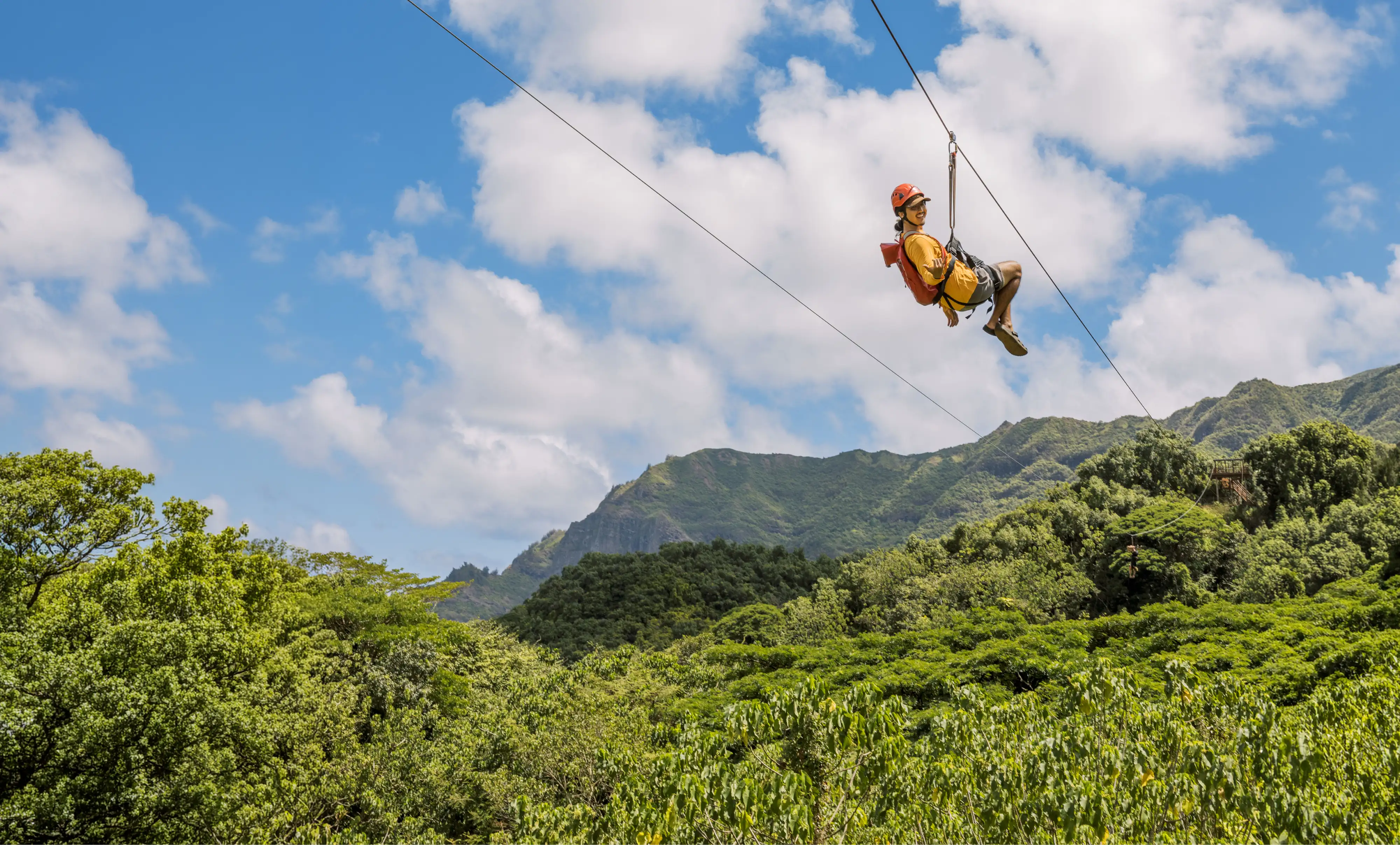 Person wearing a helmet and harness smiling while ziplining over green forest and mountains.