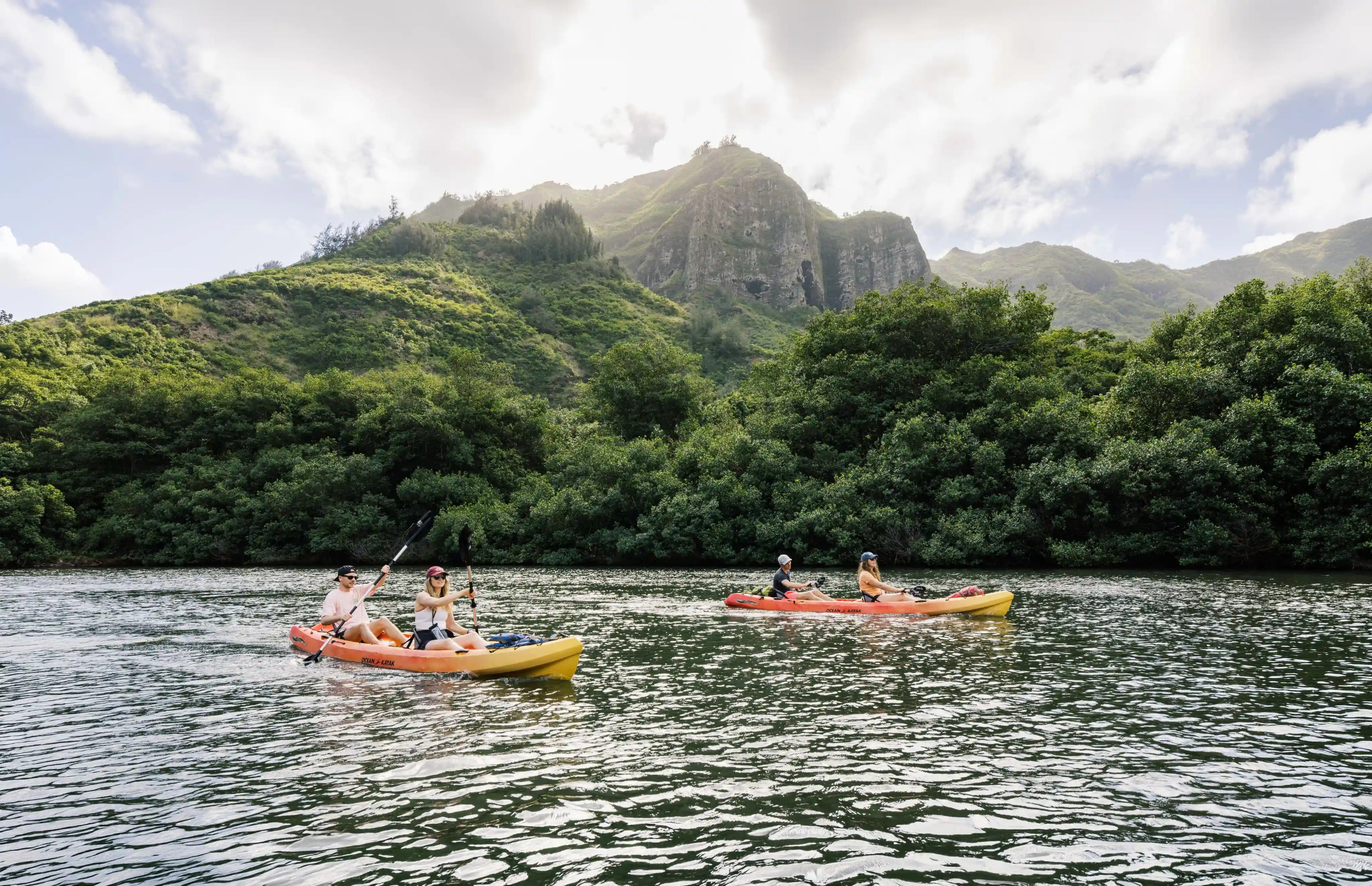 Two kayaks with four people paddling on a river with green mountains and cloudy sky in the background.