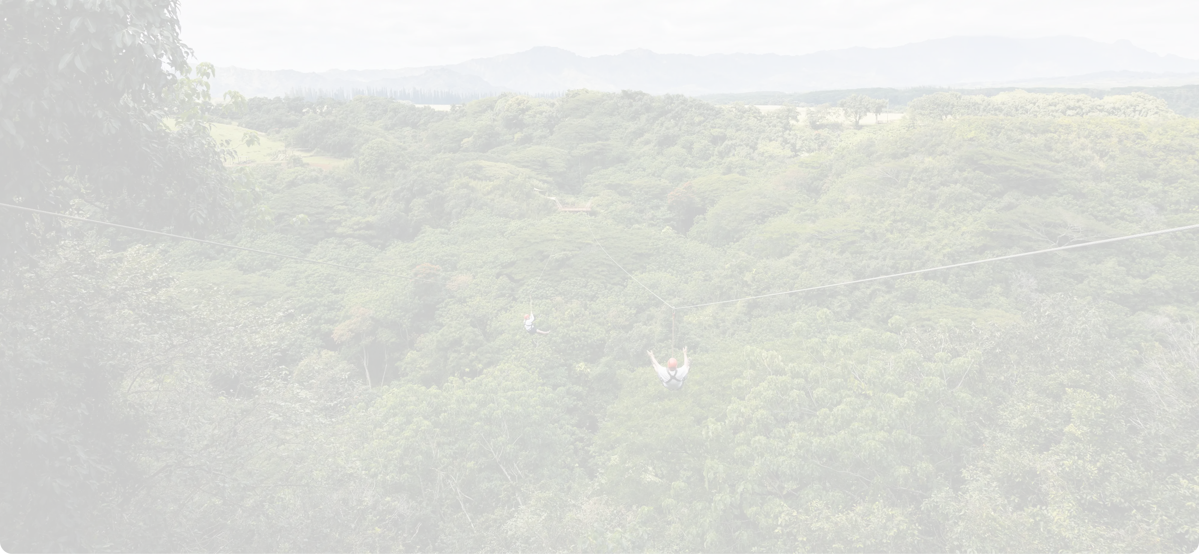 Two people ziplining over a dense green forest with mountains in the background under a cloudy sky.