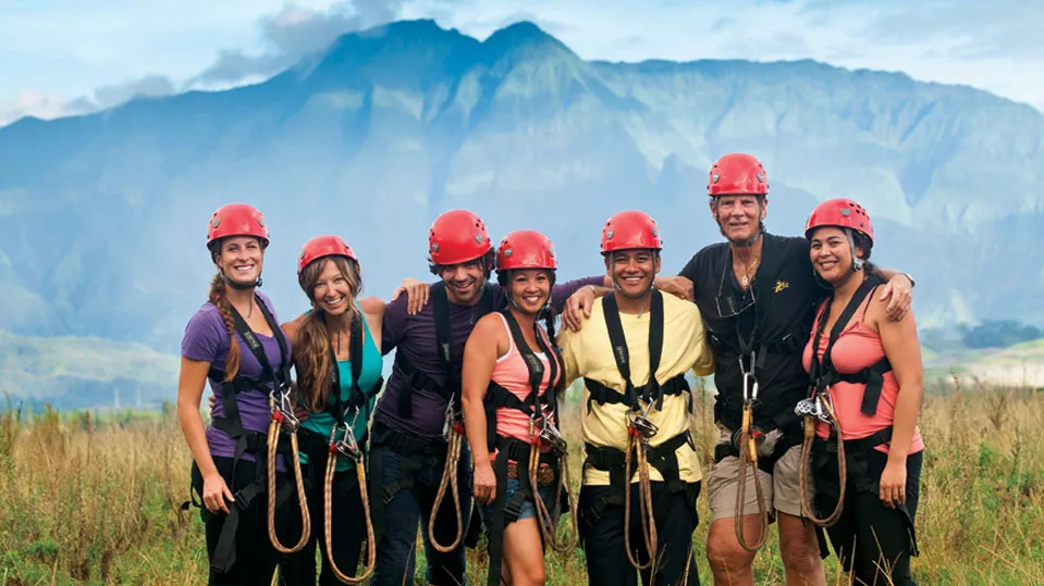 Seven smiling guests in red helmets and harnesses pose in a grassy field before green mountains.