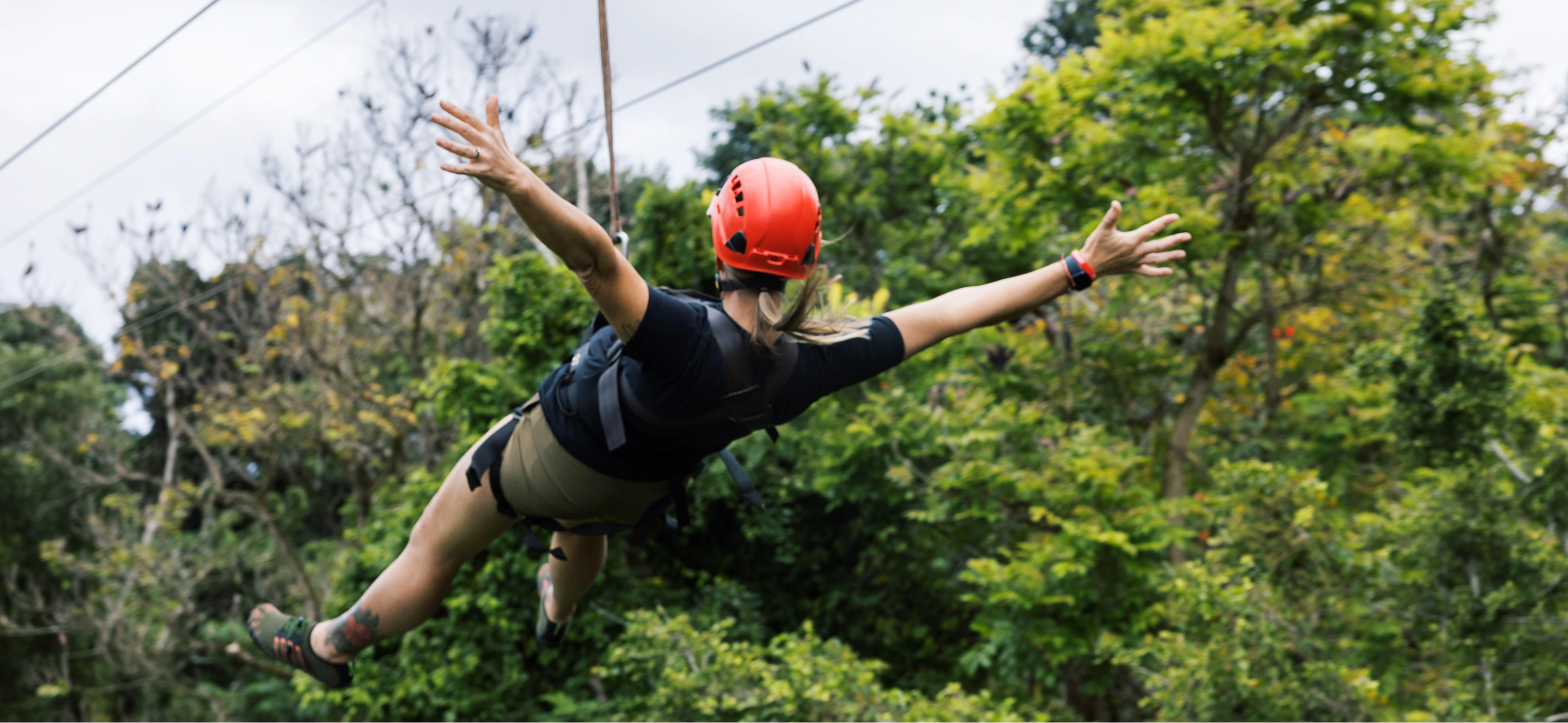 Person in an orange helmet ziplines through lush green forest with arms spread wide.