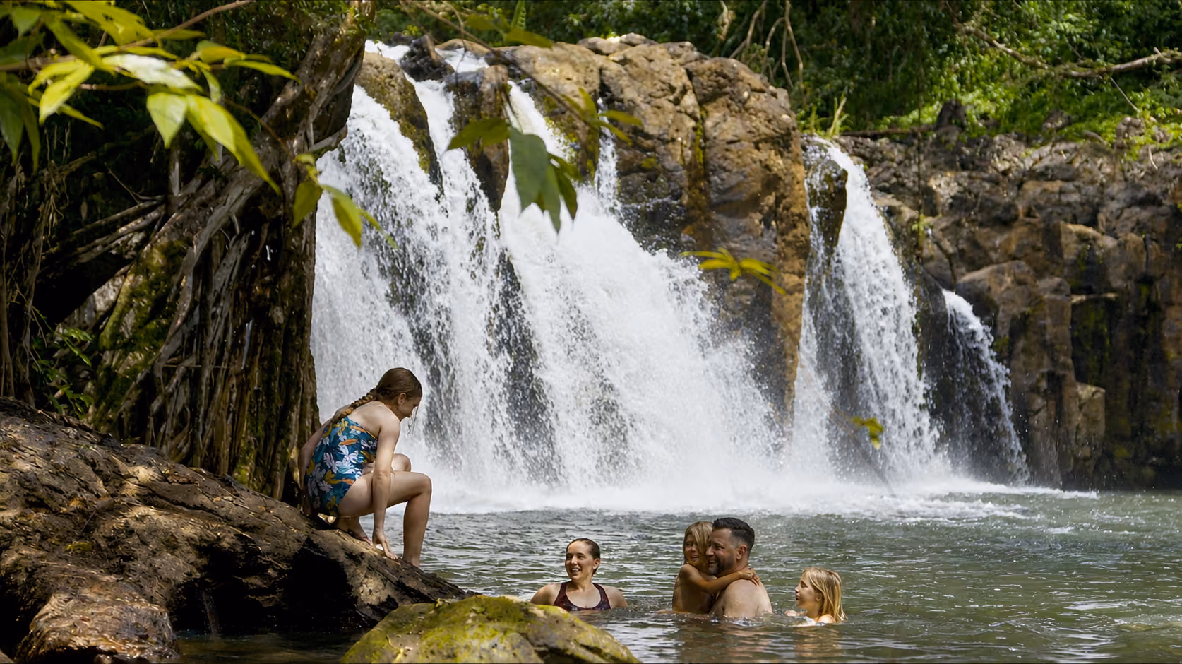 Hikers on a forest trail approaching a waterfall deep in Kauai's tropical landscape.