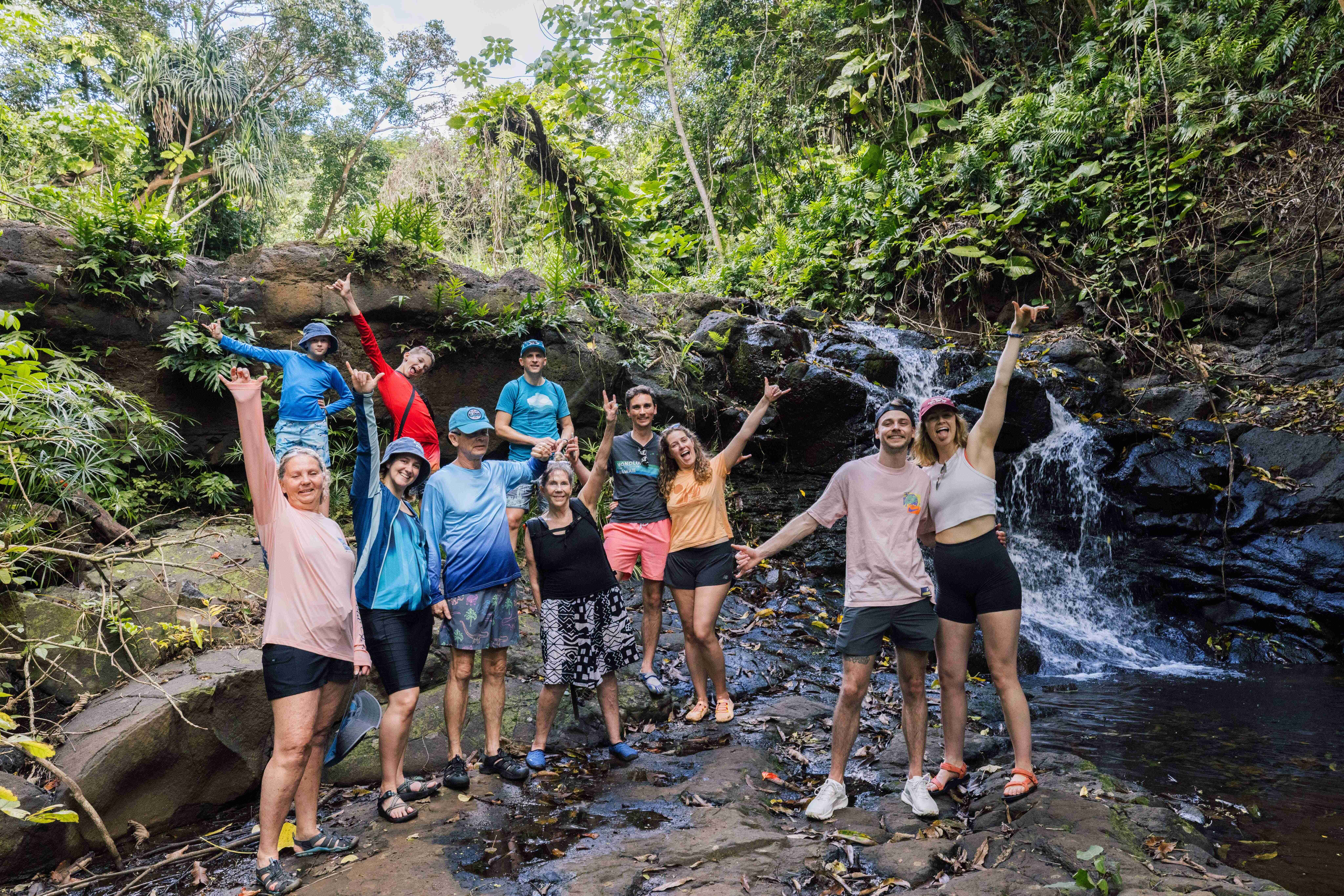Eleven travelers pose happily beside a cascading waterfall in a lush tropical jungle.