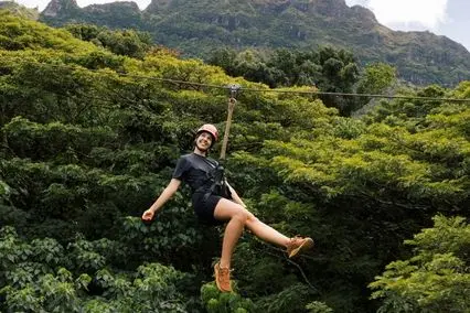 Young woman in helmet ziplining joyfully above dense green forest with mountains in background.